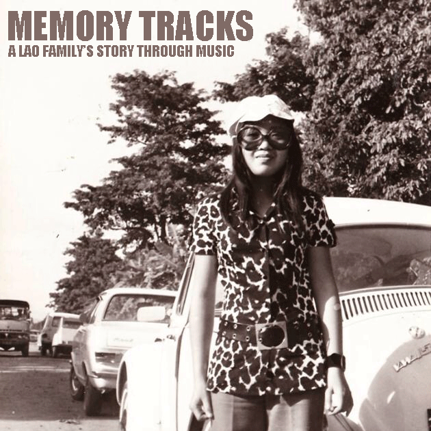 A young Asian woman wearing a hat, oversized round sunglasses, and a 1970's printed top and bell bottoms is seen standing in front of an old Volkswagon Beetle and a row of other old cars, standing on a dirt road with trees in the background.