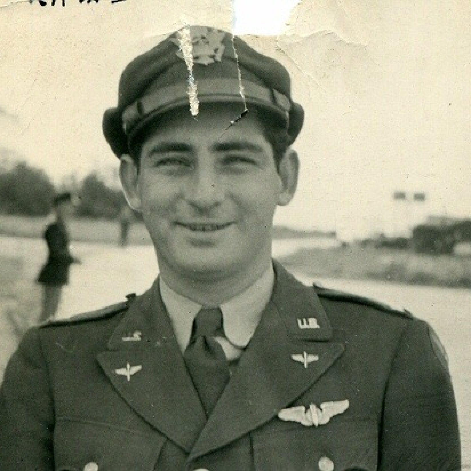 Black and white photograph that is crumpled on the top depicting a young white man smiling and wearing a military hat and uniform.