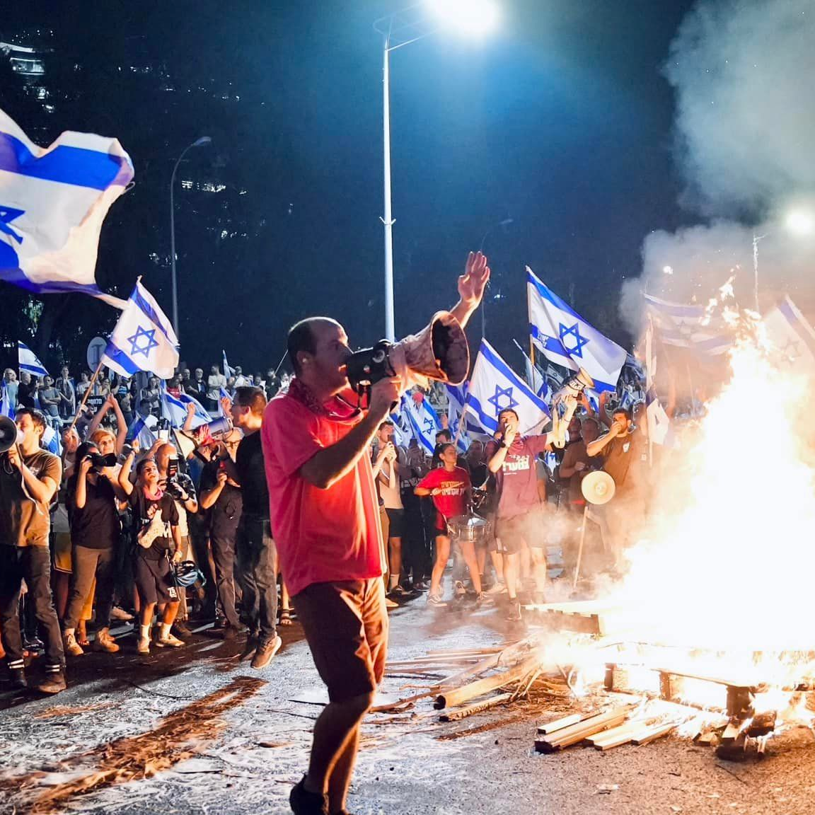 One of the many rallies of the Israeli democracy protest movement (protesters with Israel's flag gathered around a bonfire, stopping traffic on a highway). Photo: Itai Raziel.