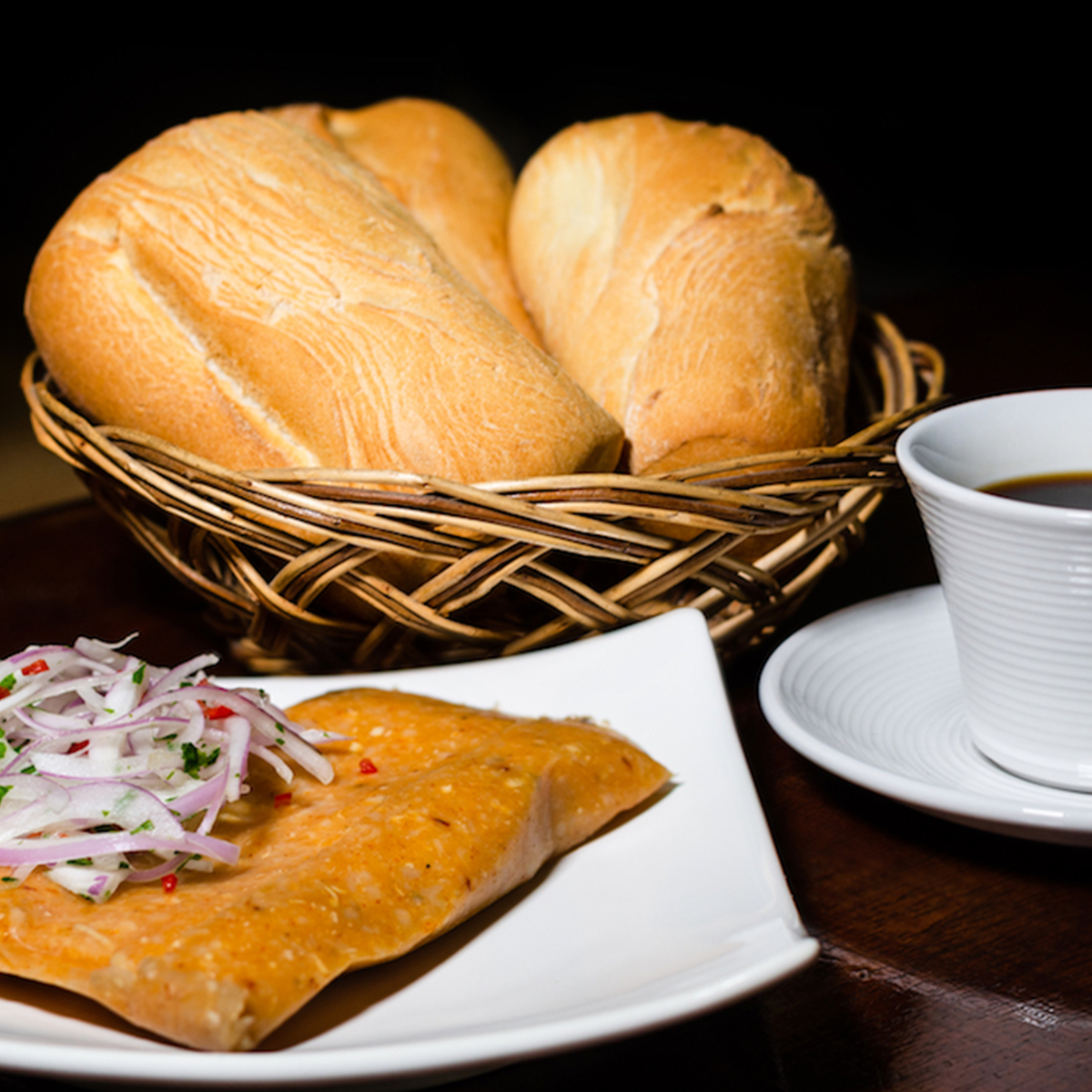 A popular Peruvian breakfast: a tamale served with salsa Criolla (pickled onions) on a square plate. Adjacent is a cup of coffee and a basket full of bread rolls. Photo Credit: ID 209102501 © Juan José Napuri Guevara | Dreamstime.com