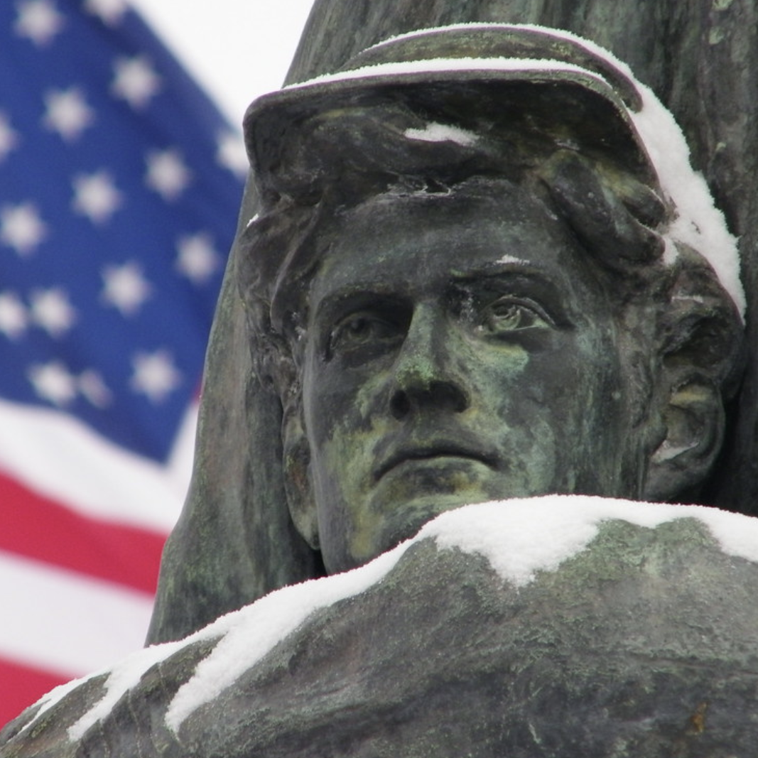 The Student Veteran Memorial pictured here was built by Chicago sculptor Lorado Taft, who built the sculpture to honor Hillsdale College students who fought and died during the American Civil War. The head of a weathered statue, a young soldier in cap and uniform, fills the foreground while a waving American flag drops out of focus in the background.